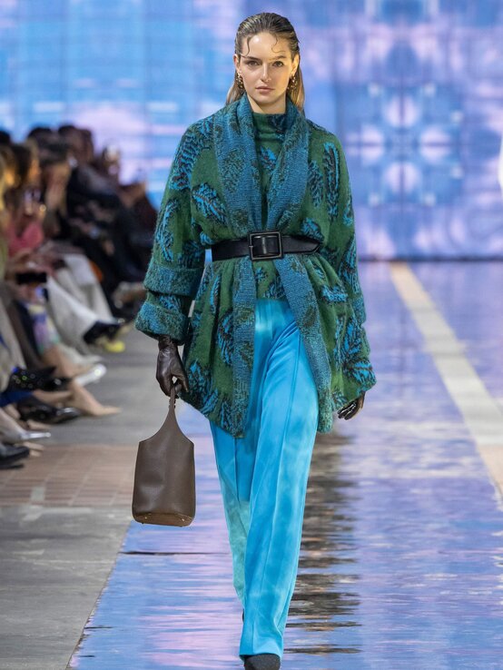 Ein Model mit zurückgekämmtem Haar läuft in einem blauen Hosenanzug und grünem Mantel mit schwarzem Gürtel über den Laufsteg, eine braune Tasche in der Hand. | © Marc Cain