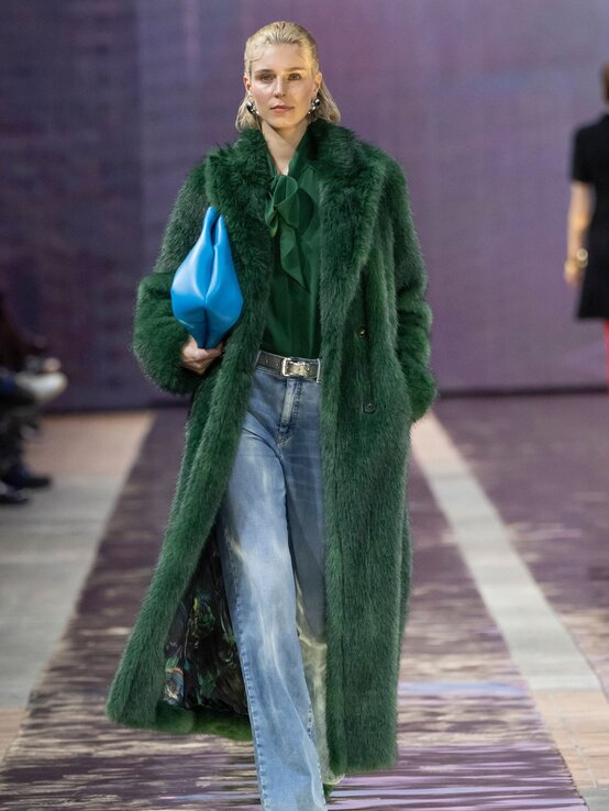 Eine blonde Frau mit zurückgekämmtem Haar trägt einen langen grünen Kunstfellmantel, eine grüne Bluse mit Schleife, weite Jeans und eine blaue Clutch. | © Marc Cain 