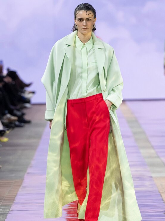 Ein Model mit strengem Blick läuft in einem mintgrünen Mantel, passender Bluse und leuchtend roter Anzughose über den Laufsteg. | © Marc Cain 