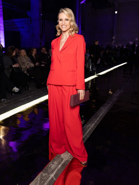 Elegant gekleidete Michelle Hunziker in rotem Anzug mit weitem Schnitt posiert mit glänzender Clutch und Sandaletten auf reflektierendem Boden. | © Andreas Rentz/Getty Images