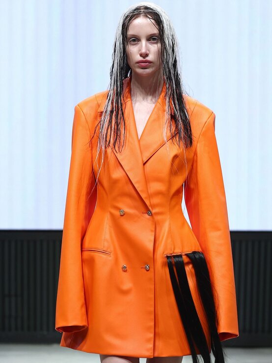 Model mit nassen, schwarz-weiß gefärbten Haaren trägt einen knallorangen Blazer mit auffälligen Haarsträhnen als Designelement auf dem Laufsteg. | © Sebastian Reuter