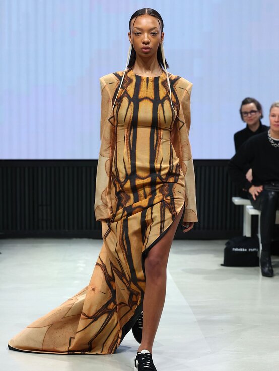 Frau in avantgardistischem Kleid mit abstraktem Muster, langen Ärmeln und Schleppe kombiniert den Look mit schwarzen Sneakers auf dem Catwalk. | © Sebastian Reuter