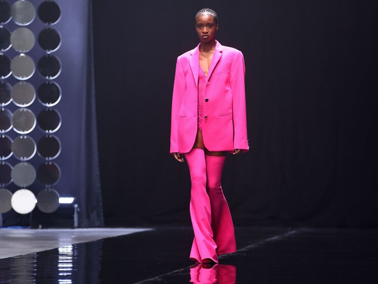 Modisches Model in leuchtendem Pink kombiniert Oversize-Blazer mit Stiefelhose und präsentiert den Look auf einem glänzenden Laufsteg. | © Joern Pollex