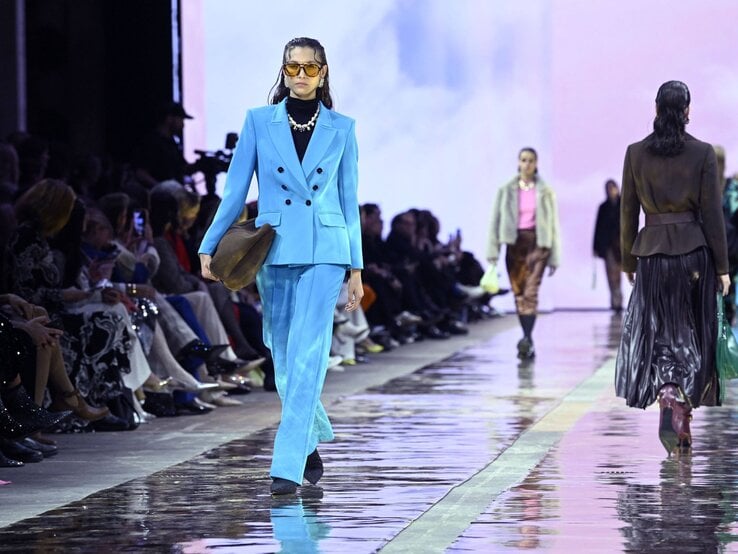 Berlin Fashion Week: Ein Model in elegantem blauem Anzug mit schwarzen Schuhen und Retro-Sonnenbrille läuft vor Publikum über den Laufsteg. | © IMAGO / Future Image