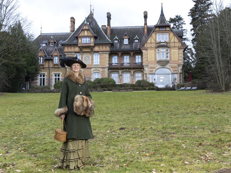 Eine elegant gekleidete Frau in grünem Mantel mit Pelzbesatz und Federhut steht mit Korb vor einem historischen Herrenhaus. | © Bernd Hanselmann