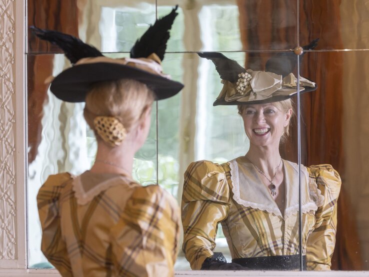 Eine Dame im historischen Kleid betrachtet sich mit einem strahlenden Lächeln im Spiegel, ihr kunstvoller Hut mit Federn im Fokus. | © Bernd Hanselmann