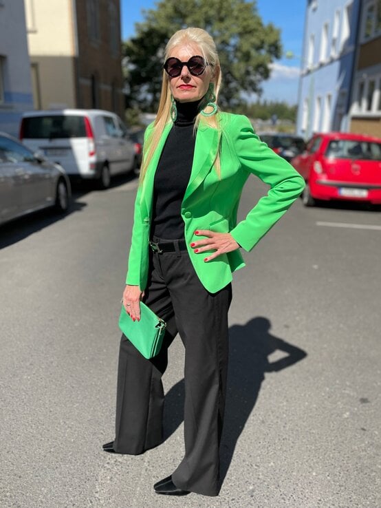 Renate Zott mit langem blondem Haar, schwarzer Sonnenbrille und grünem Blazer posiert mit Handtasche auf einer sonnigen Straße. | © Renate Zott