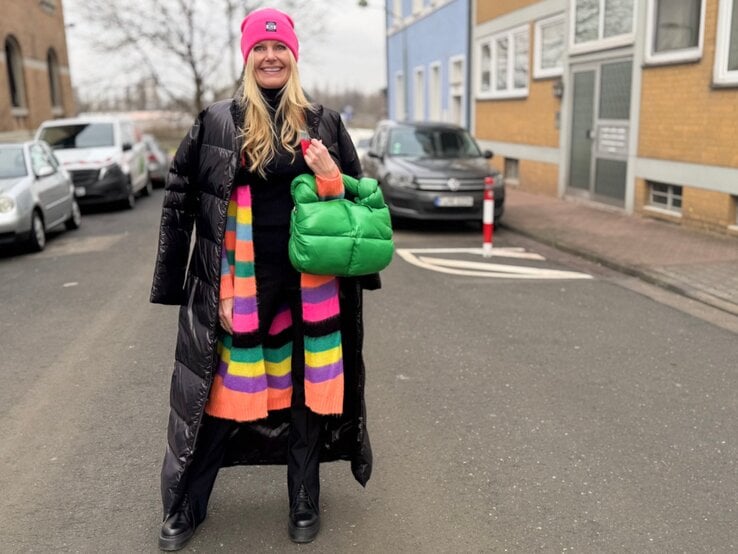 Renate Zott mit pinker Mütze, langem schwarzen Steppmantel und bunt gestreiftem Schal steht mit grüner Tasche auf einer Stadtstraße.