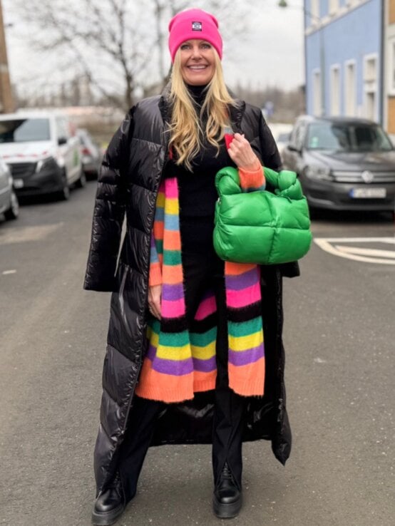 Renate Zott mit pinker Mütze, langem schwarzen Steppmantel und bunt gestreiftem Schal steht mit grüner Tasche auf einer Stadtstraße. | © Renate Zott