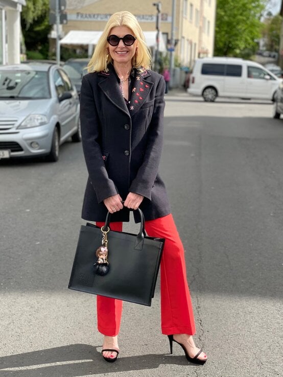 Renate Zott mit schwarzem Mantel mit Stickereien und roter Hose steht lachend auf einer Straße und hält eine große schwarze Handtasche. | © Renate Zott