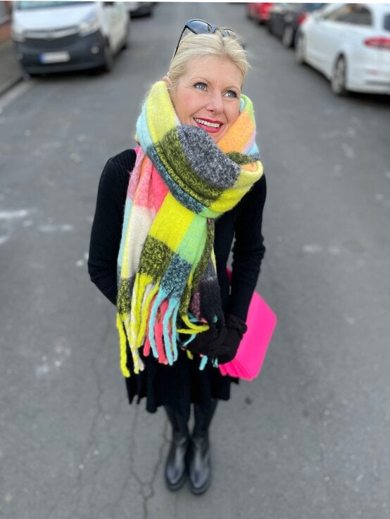 Renate Zott mit hochgestecktem Haar, buntem XXL-Schal und schwarzem Outfit steht auf einer Stadtstraße mit geparkten Autos. | © Renate Zott