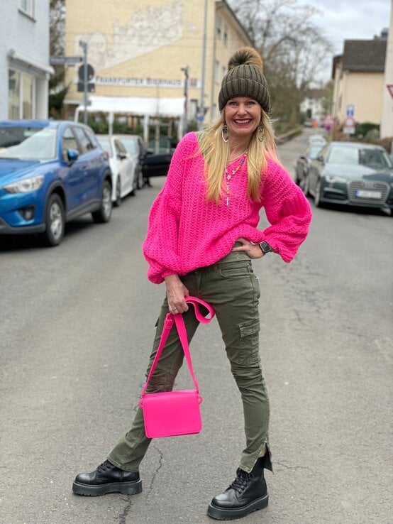 Renate Zott mit pinkem Strickpullover, olivgrüner Cargohose und Bommelmütze posiert mit einer passenden pinken Handtasche auf der Straße. | © Renate Zott