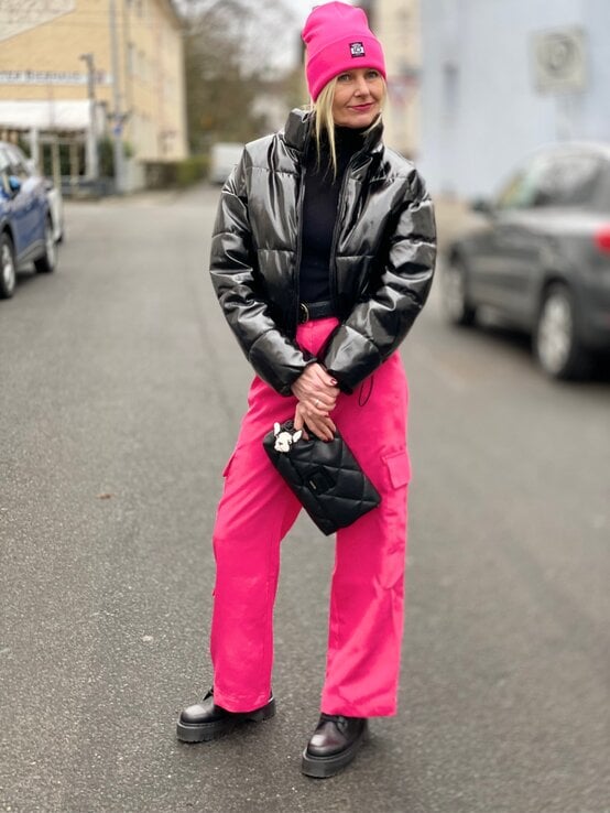 Renate Zott mit pinker Mütze, schwarzer Lackjacke und pinker Cargohose steht lächelnd auf einer Straße mit geparkten Autos. | © Renate Zott