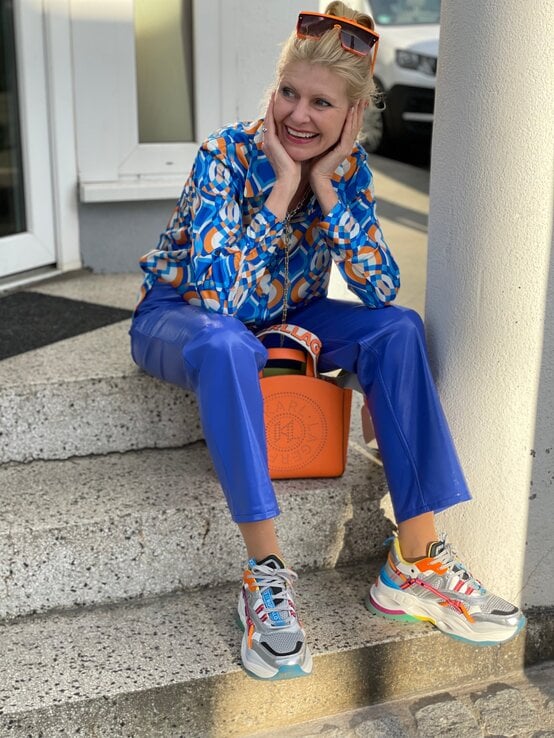 Renate Zott mit knalliger Mode in Blau und Orange sitzt auf Stufen, ihre Handtasche neben sich, während Sonnenlicht ihr Gesicht erhellt. | © Renate Zott