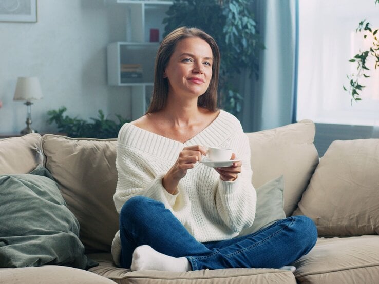 Lächelnde Frau in weißem Strickpullover und Jeans trinkt Kaffee auf einem gemütlichen Sofa in einem hellen Wohnzimmer.