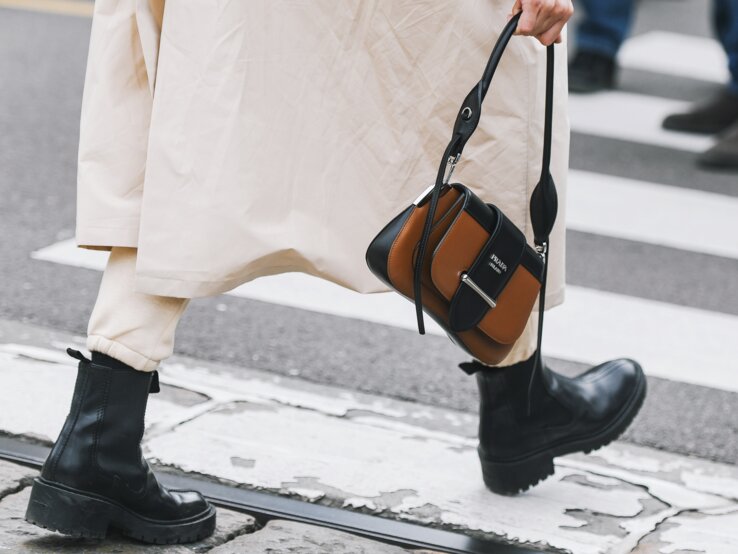 Stiefel Trends Herbst 2025 Schwarze klobige Stiefel und beige Hose unter langem Mantel, Person hält eine braun-schwarze Designerhandtasche, Gehweg im Hintergrund.