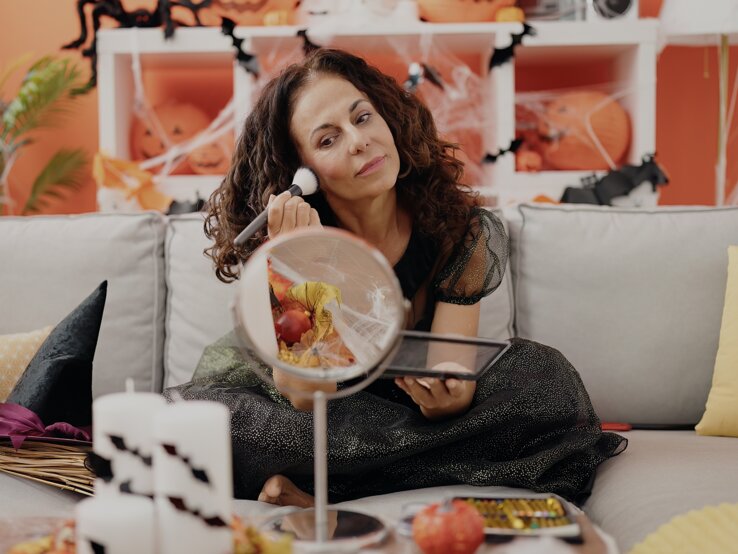 Last Minute Halloween Kostüm DIY Eine Frau in glitzerndem schwarzen Kleid sitzt auf einem grauen Sofa, mit Halloween-Dekoration und Kürbissen im orangefarbenen Hintergrund.