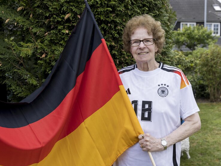 Eine ältere Frau, Oma Angelika Mittelstädt, mit lockigem Haar und Brille hält lächelnd eine große Deutschlandflagge im Garten. | © Anika Büssemeier/ BILD der FRAU