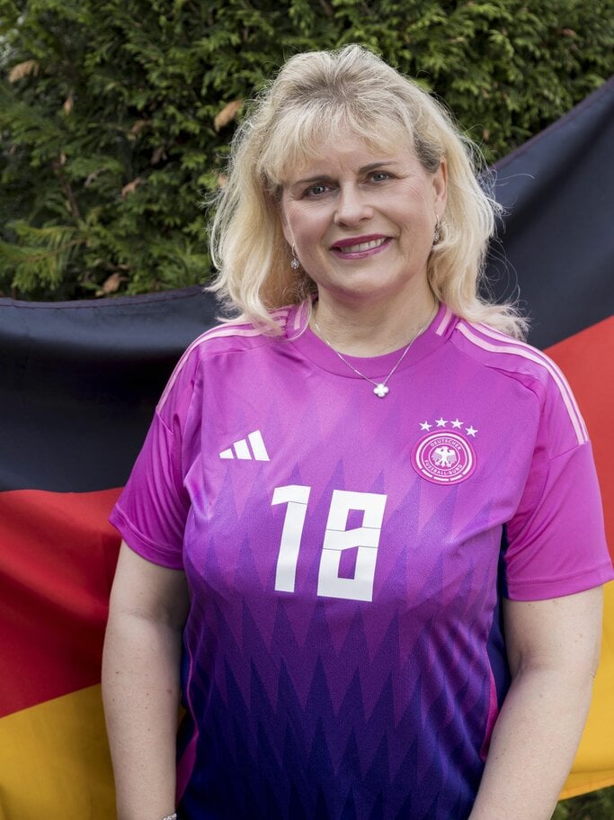 Eine lächelnde Frau im pinken Fußballtrikot der deutschen Nationalmannschaft spielt mit einem Ball vor einer dichten Hecke. | © Anika Büssemeier/ BILD der FRAU