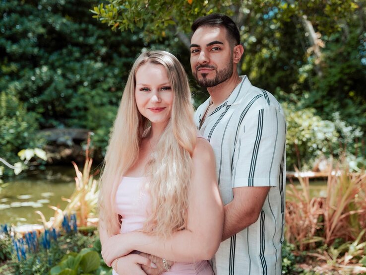 Eine blonde Frau (Sarah-Jane Wollny) und ein dunkelhaariger Mann (Tinush) posieren umarmt vor einem Teich in einem sonnigen, grünen Park.