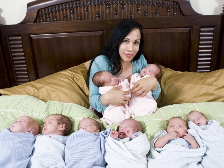 Eine dunkelhaarige Frau in einem blauen Oberteil sitzt auf einem Bett und hält zwei Neugeborene, während sechs weitere Babys in Decken vor ihr liegen.