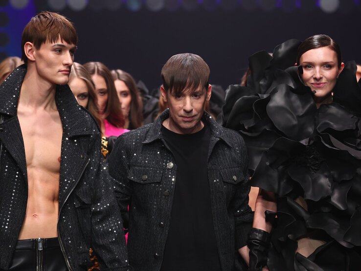 Designer Kilian Kerner mit braunem Pony schreitet mit Models in extravaganten schwarzen und glitzernden Outfits über den Laufsteg.