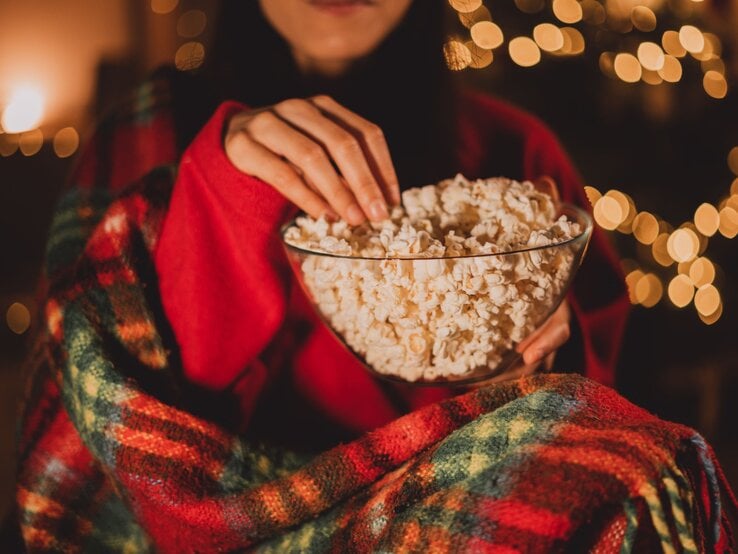 Eine Person in rotem Pullover und karierter Decke greift in eine Schüssel Popcorn vor unscharfen, warm leuchtenden Lichterketten, während sie einen Weihnachtsfilm schaut.