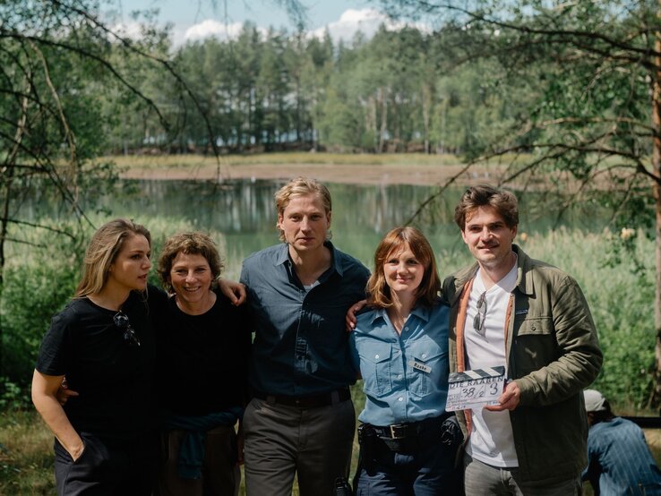 Fünfköpfiges Filmteam steht lachend vor einem idyllischen See, eine Frau in Polizeiuniform trägt ein Namensschild mit „Raabe“. | © rbb/Elliott Kreyenberg