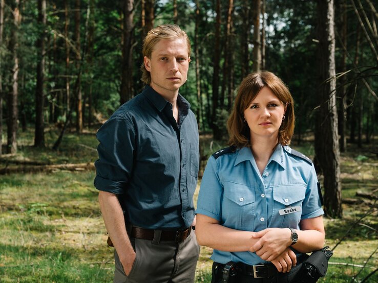 Ein blonder Mann in dunkelblauem Hemd und eine Polizistin mit braunen Haaren stehen ernst blickend in einem sonnigen Wald. | © rbb/ Elliott Kreyenberg