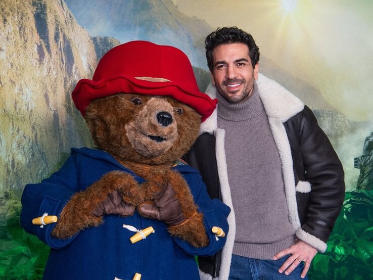 Elyas M'Barek in brauner Lederjacke mit Fellkragen posiert neben Paddington Bär, der eine rote Mütze und blauen Mantel trägt.