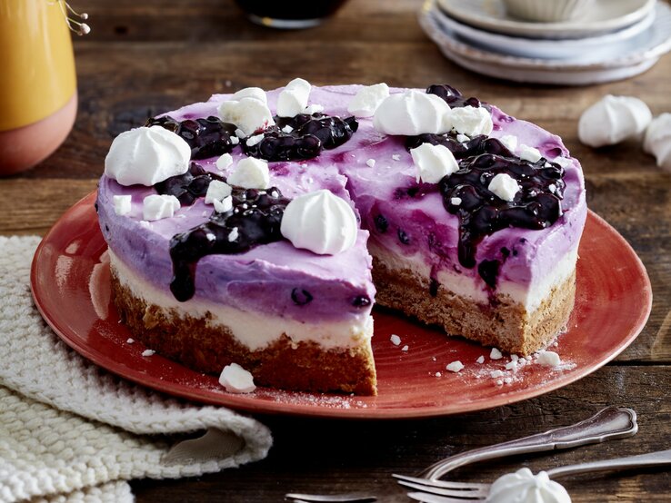 Heidelbeer-Skyr-Torte auf einem roten Teller auf hölzernem Hintergrund. Links auf dem Foto ein weißes Geschirrtuch und unten auf dem Foto Dessertgabeln. 