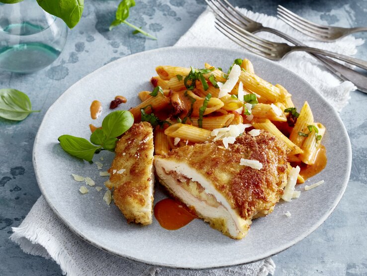 Italienisches Hähnchen-Cordon-bleu auf einem grauen Teller auf einem grauen Geschirrtuch auf grauem Hintergrund. Rechts oben drei Gabeln, links oben Wasser und Basilikum verteilt.