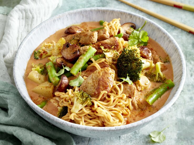 Thai-Curry mit Nudeln in einer hellgrauen Schüssel auf hellgrauem Hintergrund. Links ein Geschirrtuch, rechts Stäbchen. 