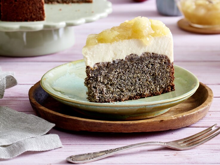 Ein Stück Joghurtcreme-Torte auf einem grünen Teller auf einem braunen Unterteller. Der Hintergrund ist lila. Rechts unten auf dem Foto eine Gabel, links unten ein Geschirrtuch. 