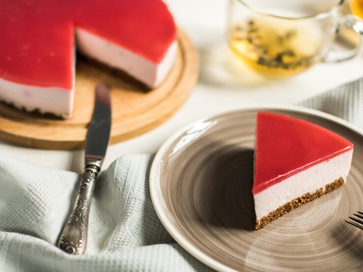 Ein Stück Schneewittchen-Torte auf einem braunen Teller. Links davon die Torte auf einem Tortenteller. Der Hintergrund ist hell. Links unten ein Geschirrtuch und rechts oben ein Tee-Glas. 