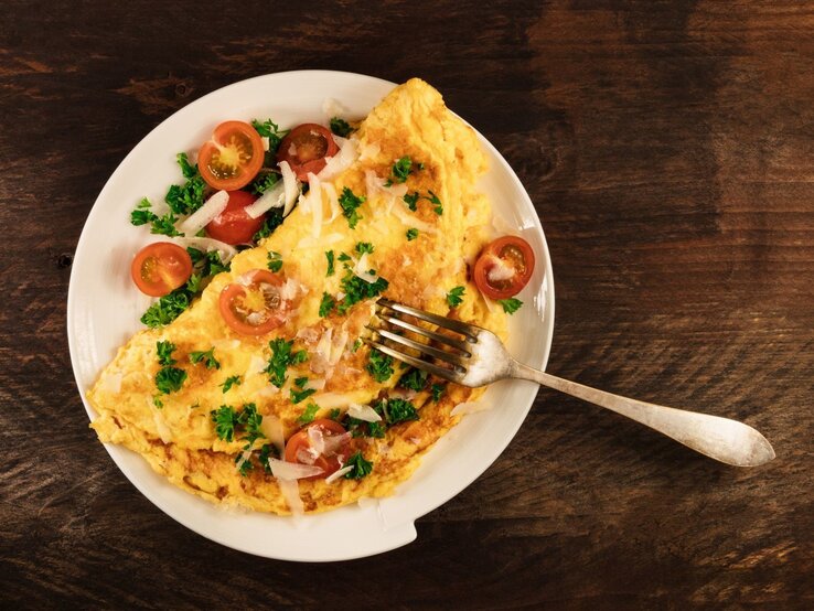 Ein goldbraunes Omelett mit Kirschtomaten, Petersilie und Parmesan auf einem weißen Teller mit einer Gabel auf dunklem Holztisch.