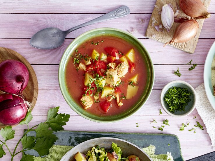 Tomatensuppe mit Huhn in einer grünen Schüssel auf rosa Hintergrund. Links davon Zwiebeln, rechts Schalotten und Petersilie. 