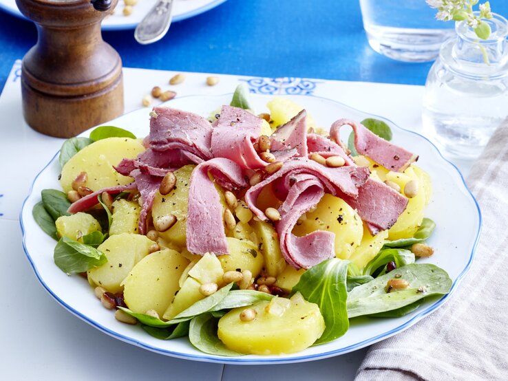 Kartoffelsalat mit Roastbeef auf einem weißen Teller auf einem weißen Tischuntersetzer auf einem blauen Tisch. Rechts ein weiß/graues Geschirrtuch und links eine Pfeffermühle.