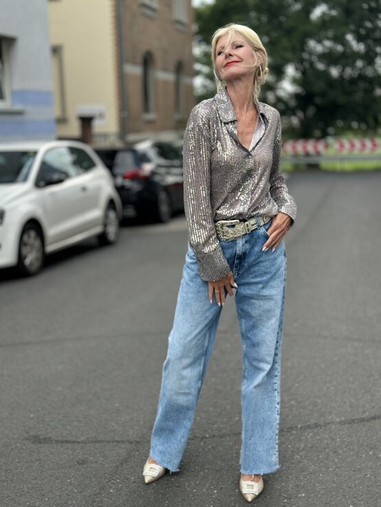 Frau mit blondem Haar, die eine funkelnde, silberne Paillettenbluse und eine hellblaue Jeans trägt. | © Renate Zott