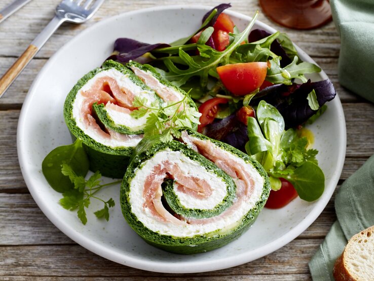 Spinat-Lachs-Roulade auf einem weißen Teller angerichtet  mit Salat auf hölzernem Hintergrund. Links davon Besteck, rechts Brot.