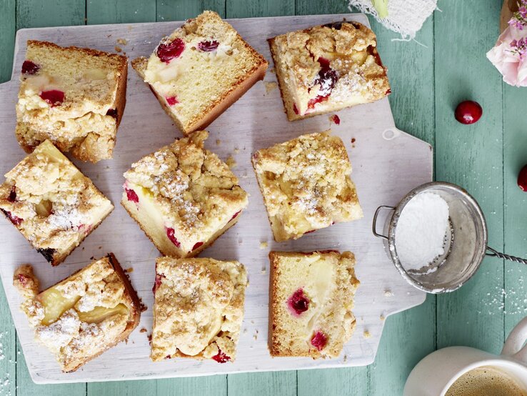 Apfel-Cranberry-Streuselkuchen auf einem weißen Schneidebrett auf grünem Hintergrund.