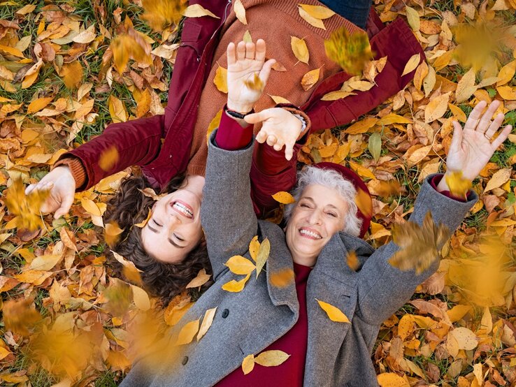 Eine ältere Frau mit grauen Haaren und eine junge Frau mit braunen Locken liegen lachend im Herbstlaub und werfen Blätter in die Luft.