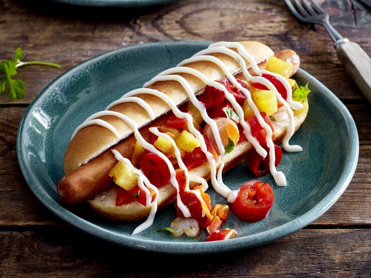 Hotdog mit Ananas & Jalapeños auf einem dunkelblauen Teller auf hölzernem Hintergrund. Rechts von dem Teller eine Gabel. 