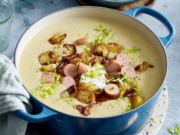 Bratkartoffel-Suppe mit Würstchen in einem blauen Topf auf grauem Hintergrund. 