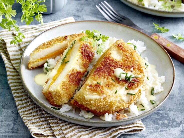 Sellerie-Cordon-bleu auf einem grauen Teller auf einem braun-weiß-gestreiften Geschirrtuch. Der Hintergrund ist grau und rechts von dem Teller liegt eine Gabel. 