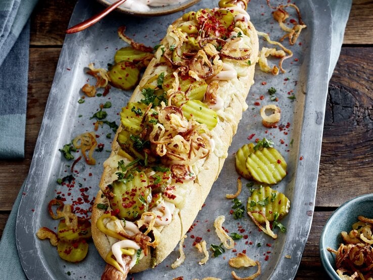 Hotdog ofengebacken im Kräuterbaguette auf einem silberfarbenen Blech auf hölzernem Hintergrund. 