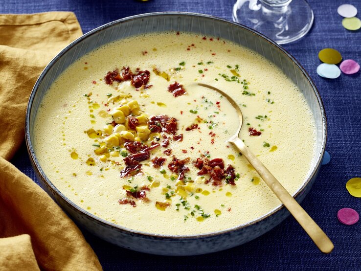 Mais-Cremesuppe in einer grauen Schüssel auf dunkelblauem Hintergrund. In der Schüssel ein goldener Löffel und links davon ein gelbes Geschirrtuch.