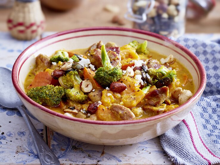 Puten-Gulasch mit Kürbis und Honignüssen in einer hellrosa Schüssel auf weiß-blauem Hintergrund. Rechts davon ein weiß-blau-kariertes Geschirrtuch und links von der Schüssel ein Löffel.