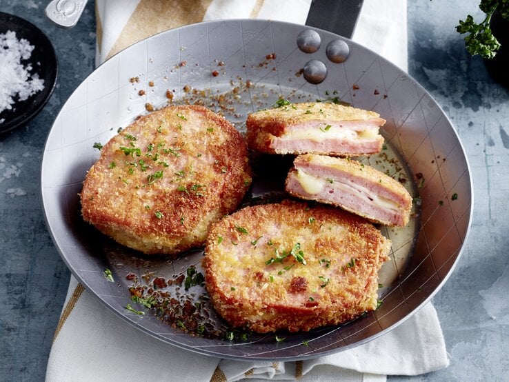 Fleischkäse-Cordon-bleu in einer Pfanne auf einem weißen Tuch auf silbernem Hintergrund.