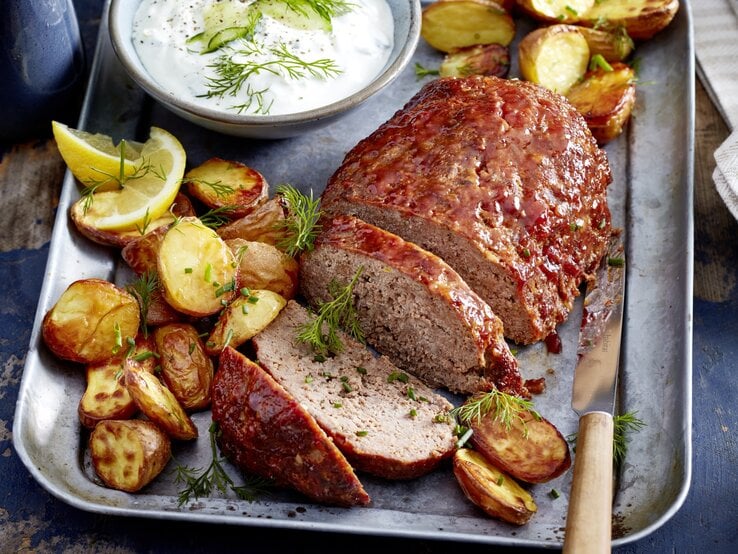 Glasierter Hackbraten mit Kartoffeln auf einem silbernen Blech. Oben auf dem Foto ein Schüsselchen mit einer Soße, rechts unten ein Messer. 